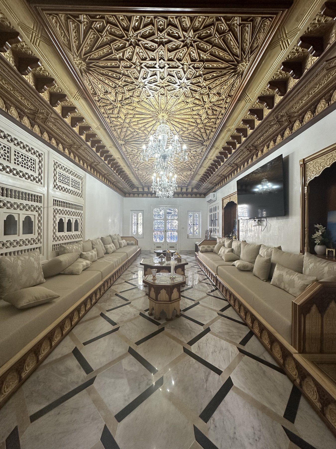 Grand salon marocain aux banquettes claires sous un plafond de bois sculpté
